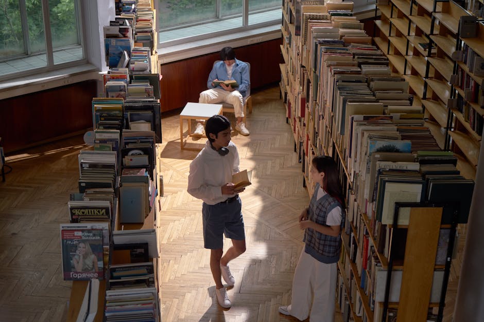 Three students in a library reading and discussing books under natural light.