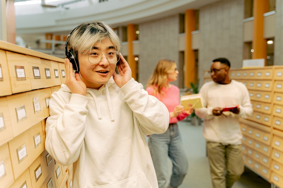 Asian man in a white hoodie listens to music with headphones in a library.