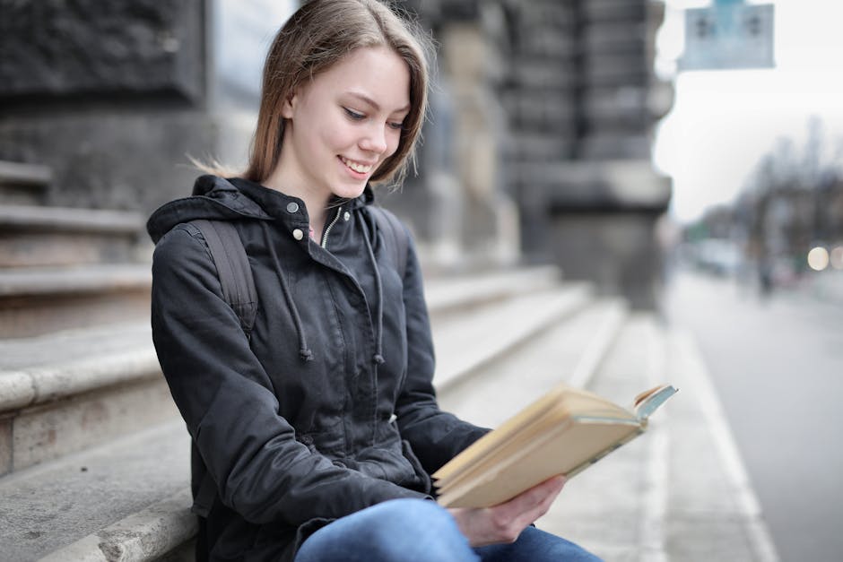 A young woman in casual attire reading a book while sitting on city steps, smiling.